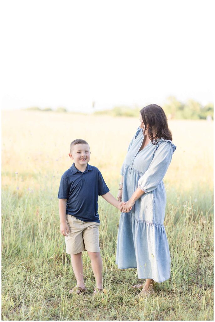 Wisp + Willow Photography Co, Texas family photographer, takes a picture of a mom holding her son's hand and looking down at him wearing a denim, long, dress and the boy smiles at the camera and wears a navy, polo, short-sleeved shirt and khaki shorts.