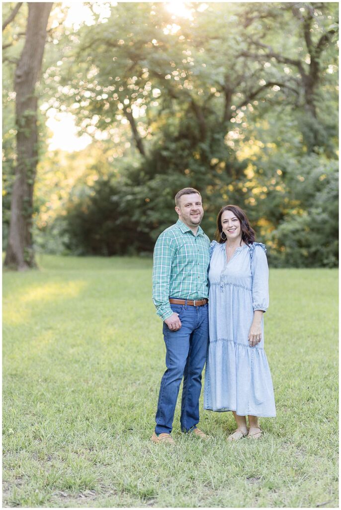 Couple stand together and smile at the camera of Wisp + Willow Photography Co. coordinating in blue and green outfits during their family portraits at Erwin Park in Texas.