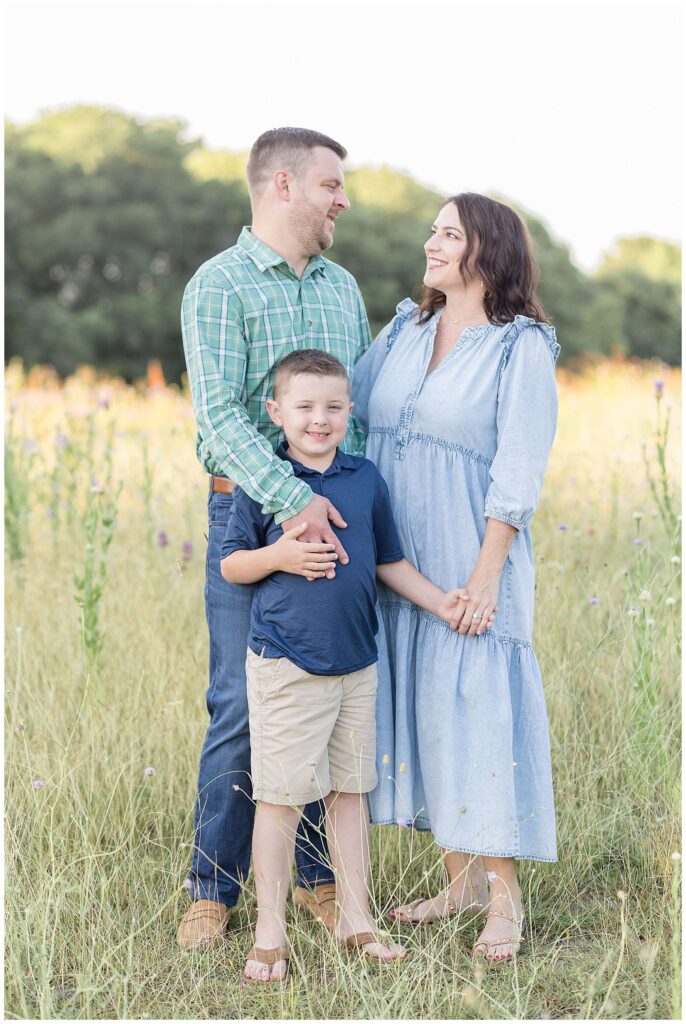 Mom and Dad look at each other as Mom holds her son's hand and Dad puts his hand on his chest as he stand in front of Mom and Dad and smiles at the camera during their Texas family portraits with Wisp + Willow Photography Co.