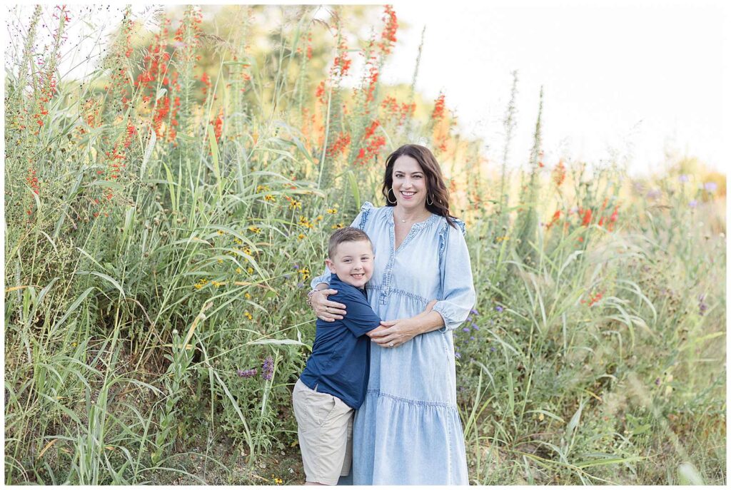 Mom and elementary-aged son, stand together as they hug and smile at the camera of Wisp + Willow Photography Co.  Mom wears a long, denim dress, and her son wears a blue, short-sleeved polo and khakis for their family portraits at Erwin Park.