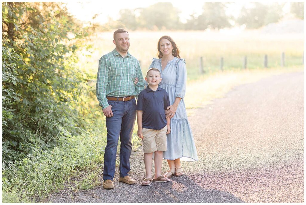 Texas family photography is gorgeous with golden hour light during family photography portraits.  This family of 3 stand with their young boy slight in front of them but standing in between as they stand on a curved, road pathway.