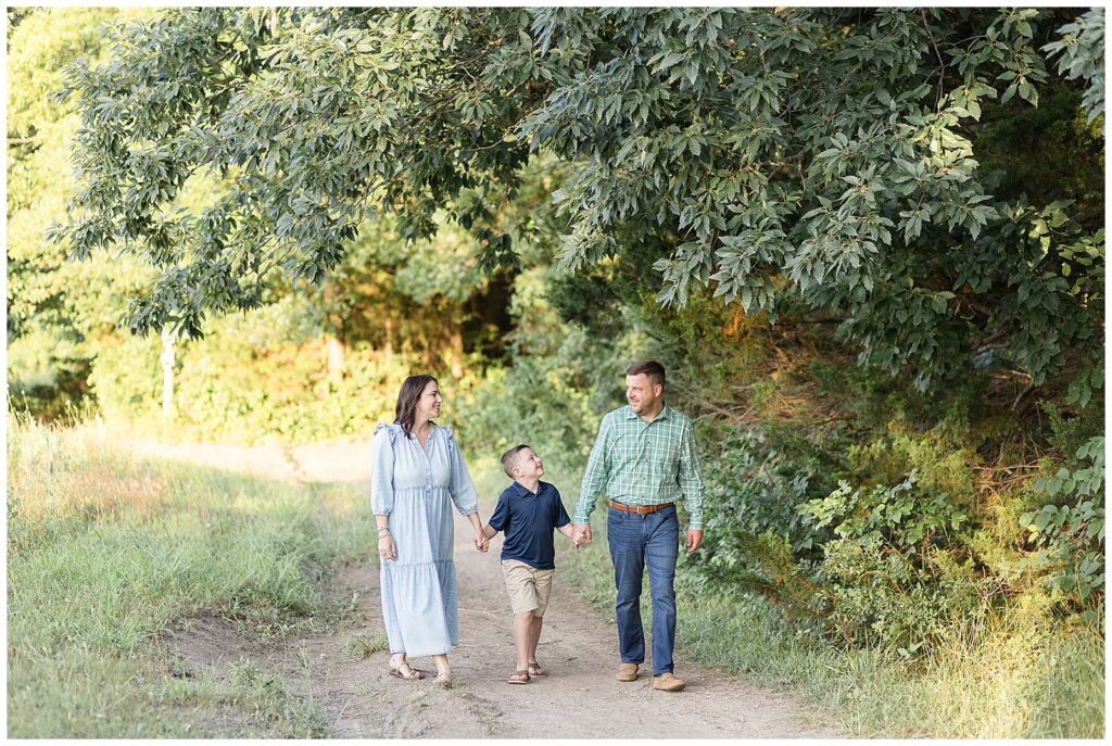 Family of 3 holding hands, walk with their young son in between his parents as they look at each and smile.  Family session at Erwin Park in McKinney, TX is taken by photographer, Wisp + Willow Photography Co.  