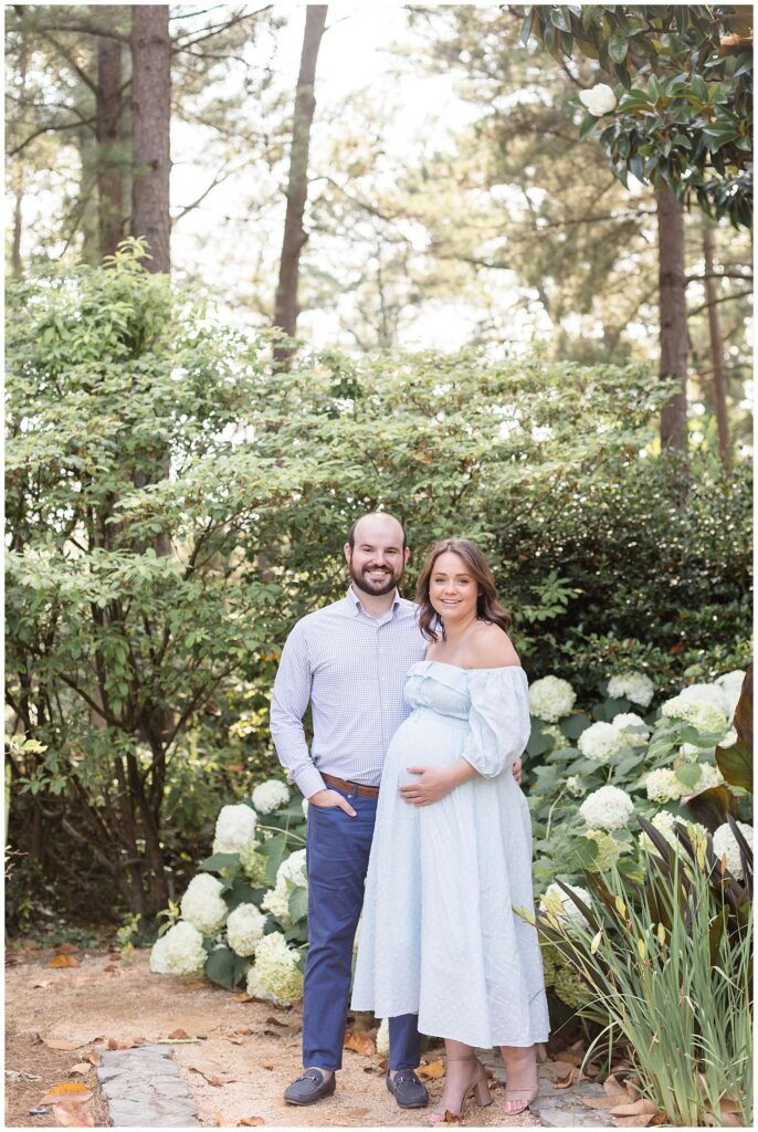 Maternity session in Raleigh with photographer, Wisp + Willow Photography Co. show an image of a couple standing in front of a hydrangea bush, surrounded with trees as they stand together and smile at the camera wearing coordinating blue outfits.