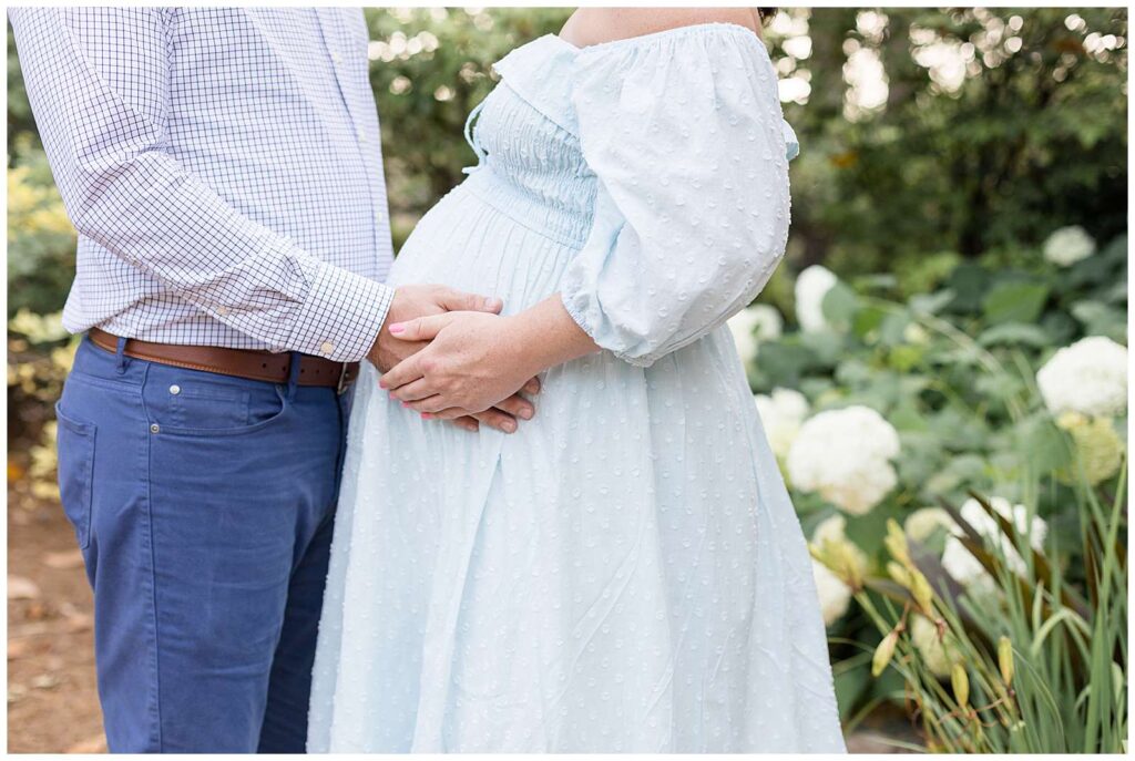Raleigh maternity portraits at WRAL Azalea Garden feature a close-up image of a couple shoulders down of the husband holding his wife's belly with her hands on top of his.