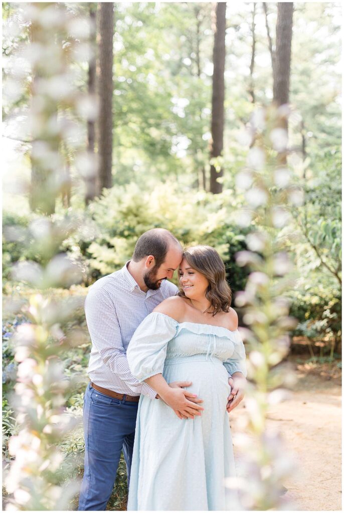 Gorgeous image of a maternity session captured by Wisp + Willow Photography Co. shows a couple through the trees of the husband giving a bear hug behind his wife and holding her belly.
