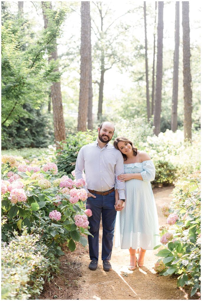 Maternity session at WRAL Azalea Gardens in Raleigh show a couple standing side by side holding hands and the wife resting her head on her husbands shoulder.  Pregnant momma wears an off the shoulder, long, mint dress with nude heels and her husband wears jeans, a blue and white small plaid, button-up shirt, and navy shoes.