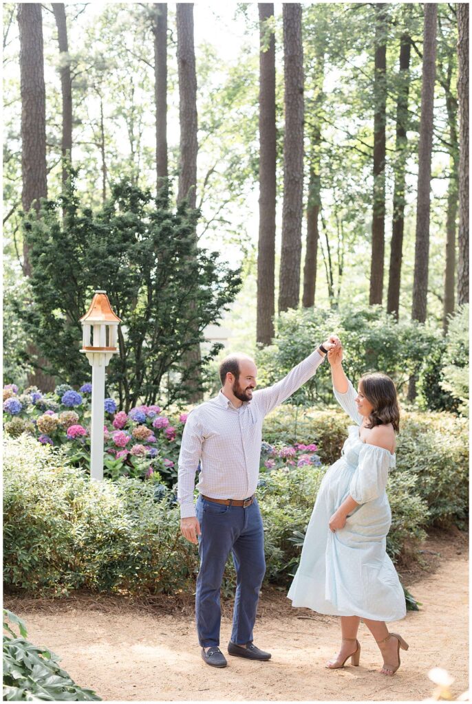 Maternity session in Raleigh, NC shows a couple dancing in the gardens at WRAL Azalea gardens.  Husband twirls wife, who is wearing a mint, long dress and they look and smile at each other.