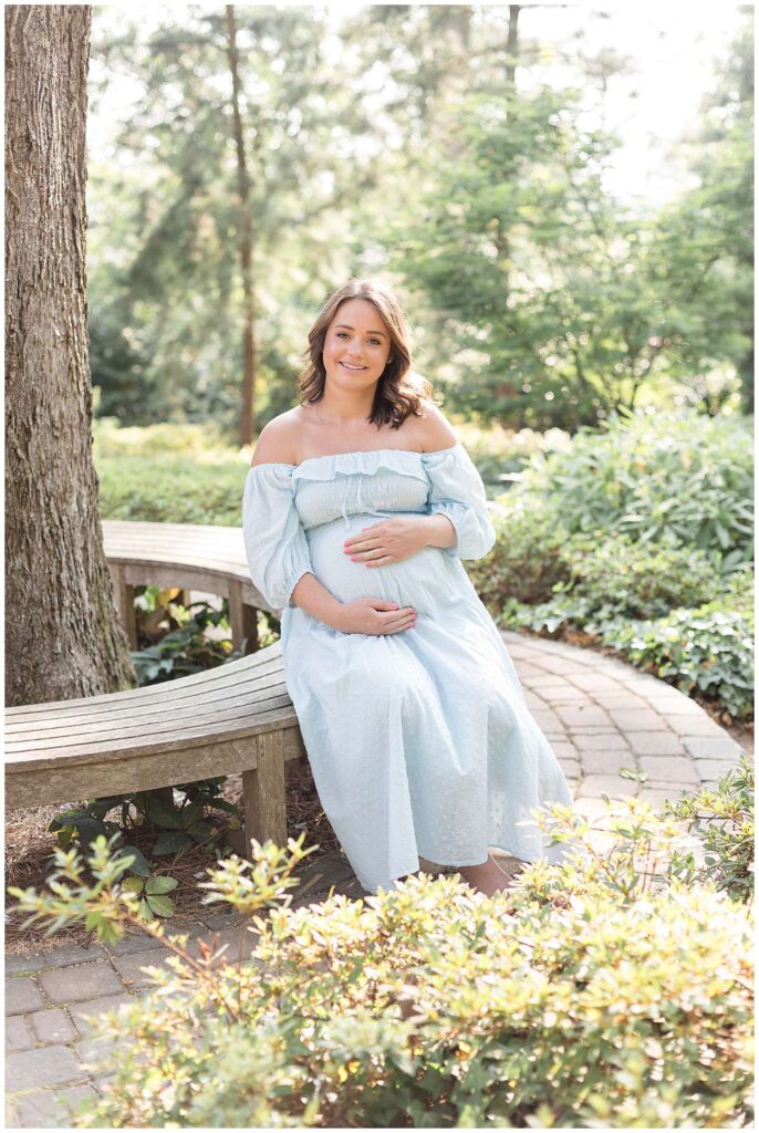 Pregnant momma-to-be sits on a circular bench surrounding a tree and she smiles at the camera of Wisp + Willow Photography Co wearing an off the shoulder, long, mint dress and holds her belly.