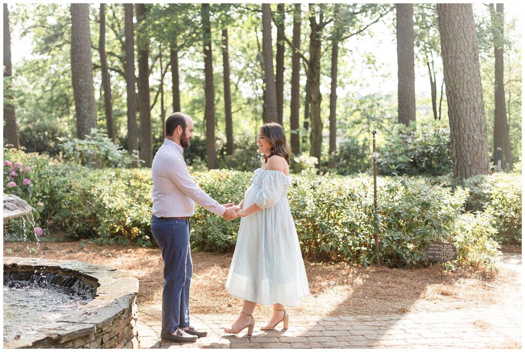 Couple holds hands and looks at each other during their Raleigh maternity session at WRAL Azalea Gardens.  The glow of the sun beams through the trees behind them as they stand near a water fountain.