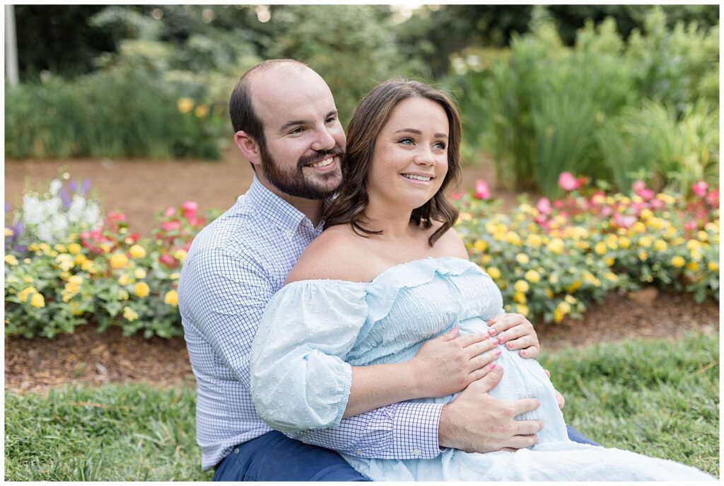 Couple sits in the grass at WRAL Azalea Gardens as pregnant mom-to-be sits back in her husbands lap and they hold her pregnant belly as they look off in the distance.