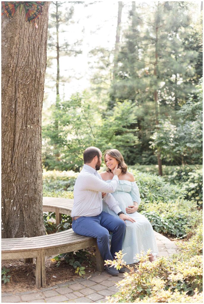 Couple sits on a circular bench facing each other as the husband pulls his pregnant wife's chin towards him for a future kiss.  They wear mint, and blue colors for their maternity session with Raleigh photographer, Wisp + Willow Photography Co.