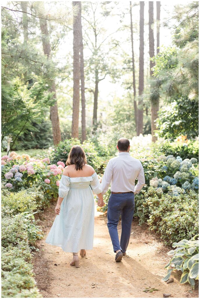 Raleigh photographer, Wisp + Willow Photography Co. take an image of a couple who walks away, hand in hand, from the camera with gorgeous sunlight coming in through the surrounding trees and with hydrangeas bordering the walkway.