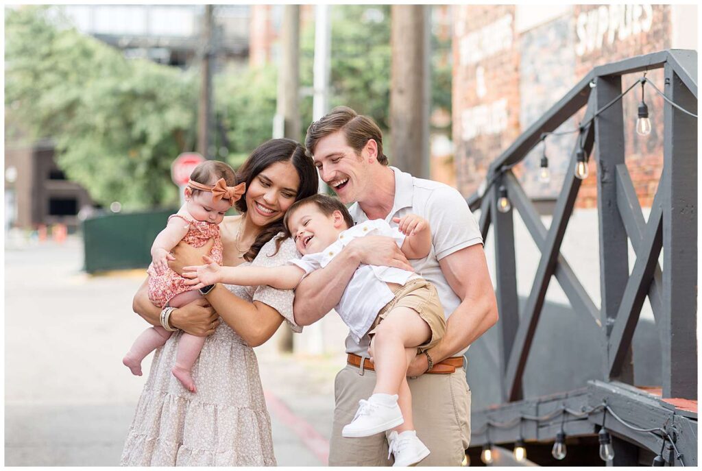 Aunt and Uncle hold their toddler nephew and baby niece as big brother reaches for his sister's hand.  They coordinate wearing neutral colors and baby girl wears a pop of muted pink during their McKinney family session.