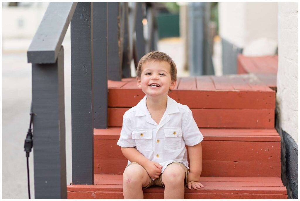 Toddler boy sits on the red steps in Downtown Square in McKinney, TX and smiles big at the camera of Wisp + Willow Photography Co. during their McKinney family session.