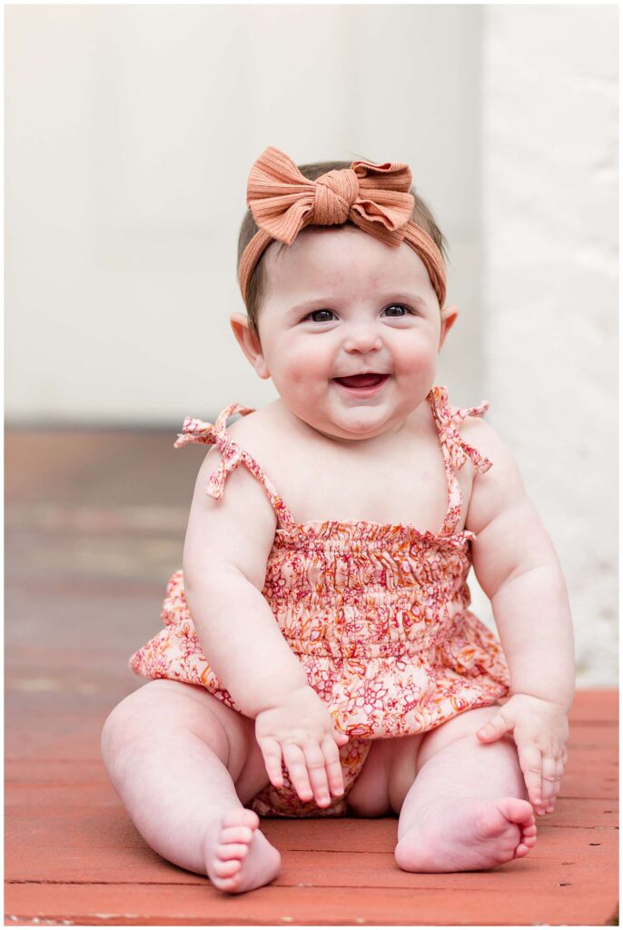 A 6 month old baby girl sits up wearing a spaghetti strap romper which a mix of pink and orange tones during a McKinney, TX family session.