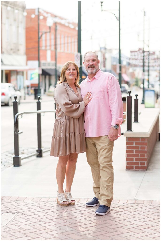 A husband and wife in their 50's or 60's stand together in Downtown Square in McKinney, TX for family portraits.  Mom wears a silk, light brown dress, and Dad wears khakis and a pink shirt as they smile at the camera of Wisp + Willow Photography Co.