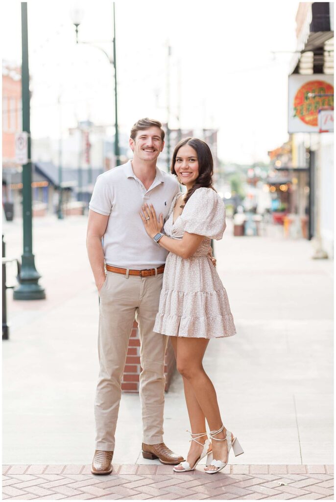 Couple stands on the sidewalk in Downtown Square in McKinney, TX with the girl wearing a khaki dress with white specks and her husband wears a cream colored polo and khaki pants.  The perfect coordinating outfits for a family session.