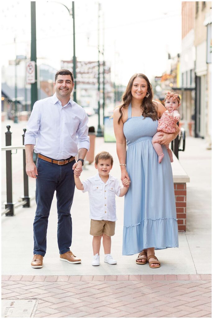 A Mom and Dad stand together holding the hands of their toddler son in between them and Mom holding her baby girl on her hip.  Mom wears a blue dress, baby girl wears a spring colored romper with color tones of pink and orange, toddler son wears a white button down shirt and khakis and Dad wears a light blue button down shirt and navy dress pants.  They stand in Downtown Square in McKinney, TX with photographer, Wisp + Willow Photography Co.