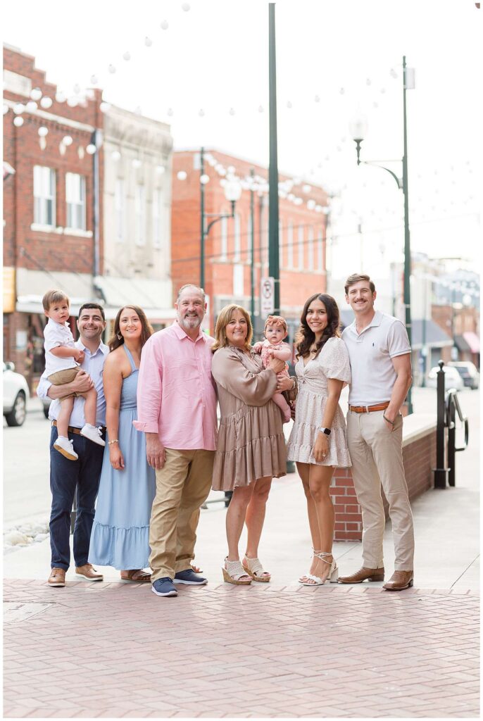 Extended family portraits consisting of 6 adults and two kids in Downtown Square in McKinney, TX have family standing on the sidewalk side by side, all coordinating in neutral colors with a pop of pink and blue for their family session.