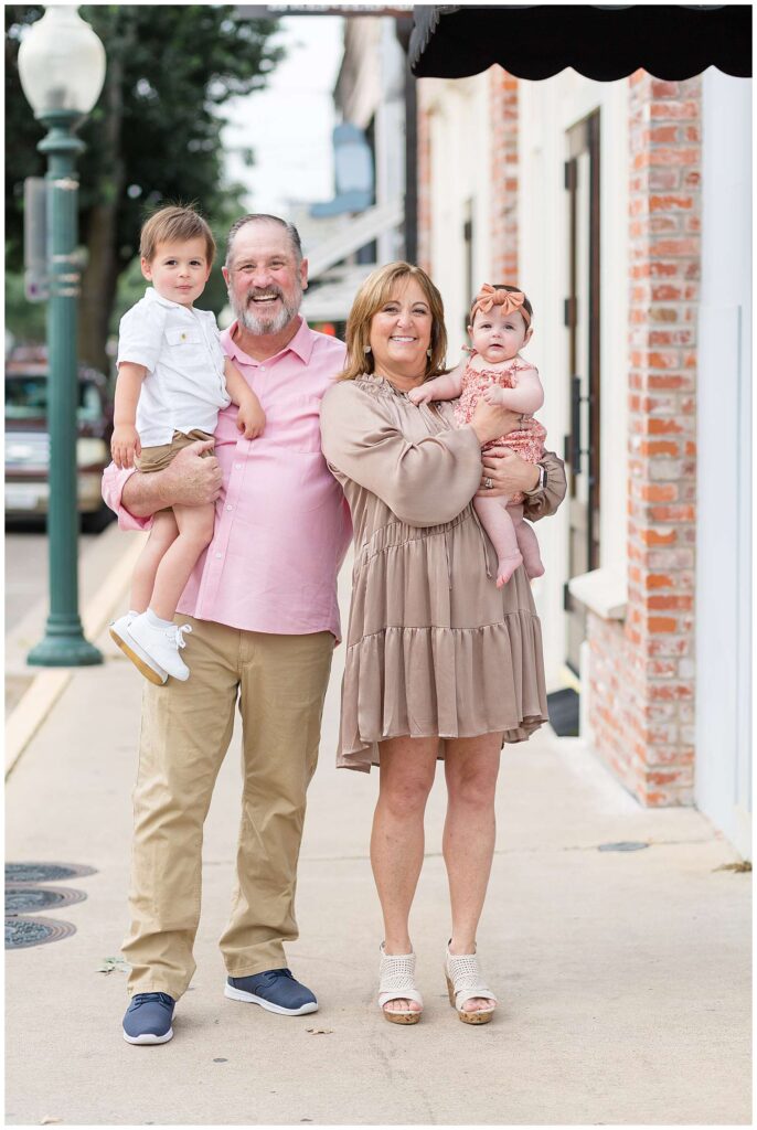Grandma and Grandpa stand on the sidewalk in Downtown Square in McKinney, TX holding their grandson and granddaughter during their family portraits with Wisp + Willow Photography Co.  They coordinate in tan colors with pops of pink and muted orange.