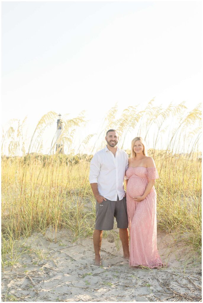 Maternity Tybee Island Beach session with Wisp + Willow Photography Co. feature a couple that stands on the beach, surrounded by sea grass and the lighthouse behind them.  Woman wears a pink, lace dress and husband wears a white button down shirt and grey shorts.