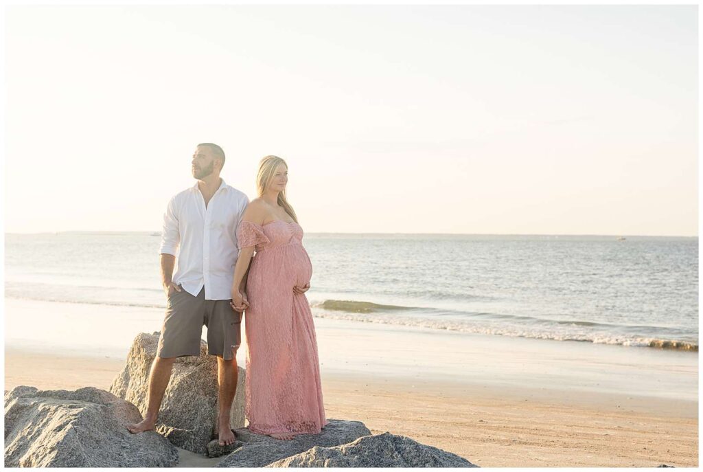 Maternity session at Tybee Island Beach in Georgia feature a couple standing on rocks with the beach behind them at sunset.  They hold hands and mom-to-be looks off in the distance as she holds her belly wearing a pink, lace dress and her husband wears a white button down shirt and grey shorts looking off in the other distance.