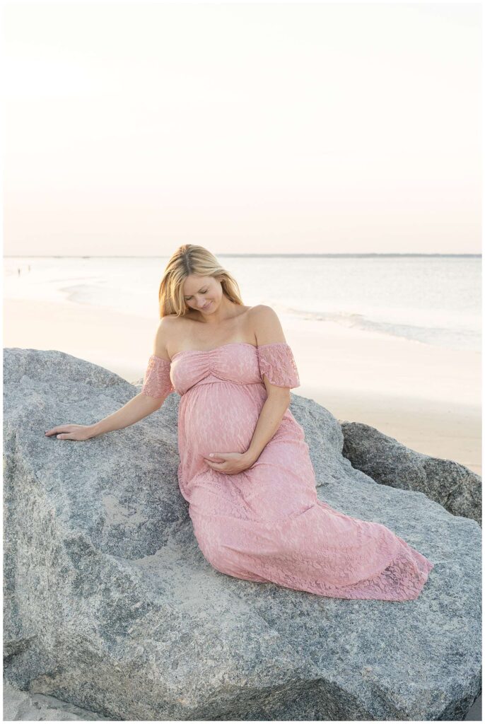 Wisp + Willow Photography Co. take a picture of a mom-to-be wearing a pink, lace dress as she sits on the rocks with the beach behind her during her maternity session at Tybee Island Beach.