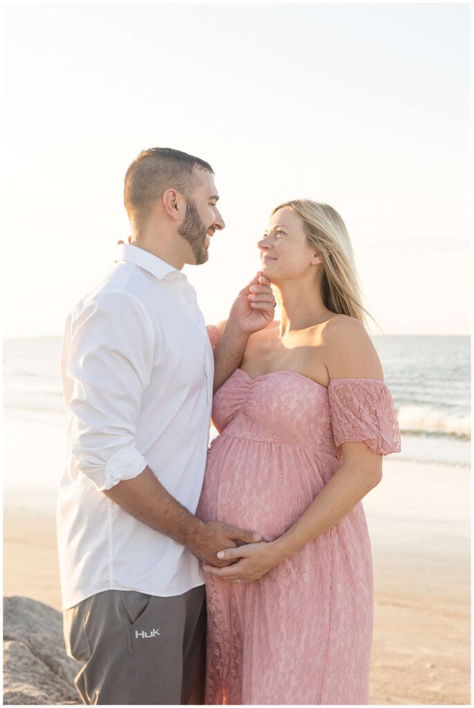 Image shows an expecting couple standing on rocks with the beach in the background.  Mom-to-be wears a pink lace dress and stands belly to belly with her husband who wears a white button shirt and holds his wife's belly with one hand and her chin with the other as he looks at her.