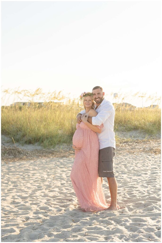 Maternity session at Tybee Island Beach shows a couple standing on the sand, surrounded with sea grass behind them as the husband gives his wife a bear hug from behind and they come cheek to cheek and smile at the camera.