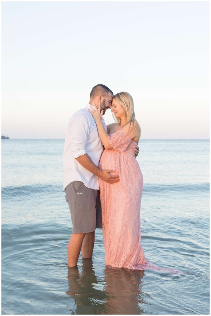 Couple stands in the ocean water belly to belly as they pull each other in close, forehead to forehead and enjoy these last couple weeks as they welcome their baby.  Pregnant woman wears a pink, lace dress and her husband wears a white button down shirt and grey shorts.