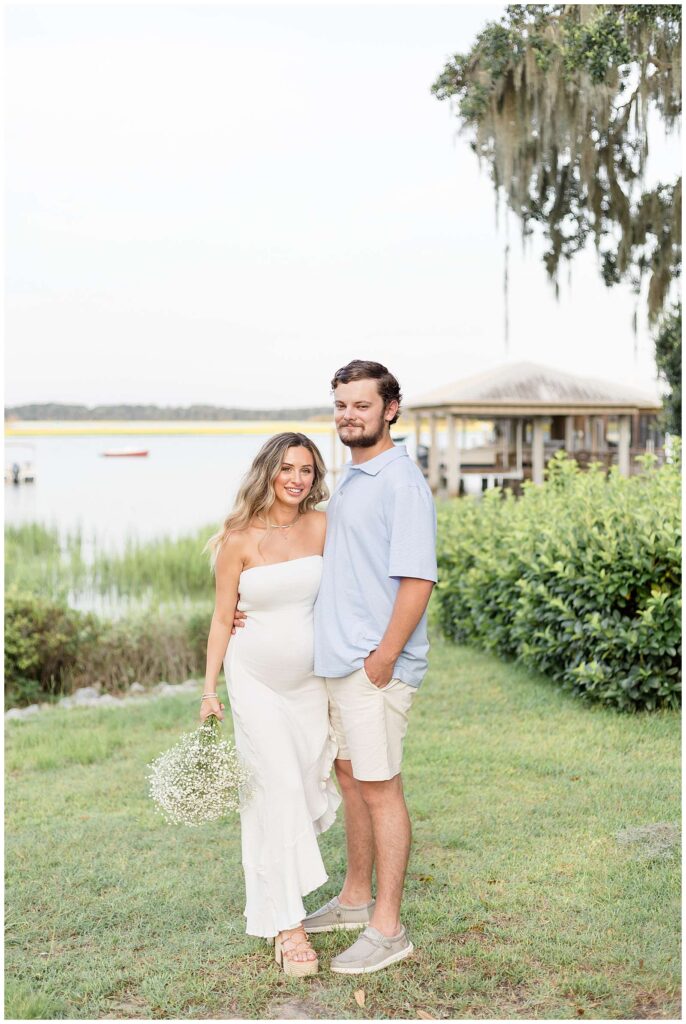 Maternity session at the Isle of Hope Marina in Savannah, GA features expecting couple who stand on the grass, with the water behind them as she wears a strapless, white dress and holds a bouquet of baby's breath in her hand and he wears a light, blue polo shirt and cream colored shorts.