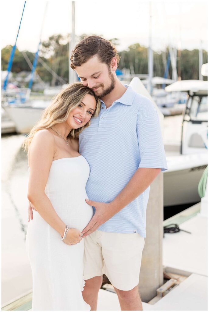 Couple stands on the dock at the Isle of Hope Marina in Savannah, GA as pregnant mom-to-be holds her belly and leans in and rests her head on her husbands chest who is looking down at her and smiling while holding her belly with their baby on the way.