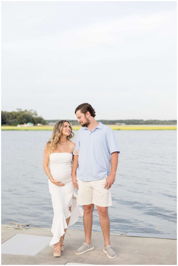 Expecting couple stand on a dock with water surrounding them as the come close, side by side, and look at each other and smile thinking about the arrival of their baby on the way during their Savannah maternity session.
