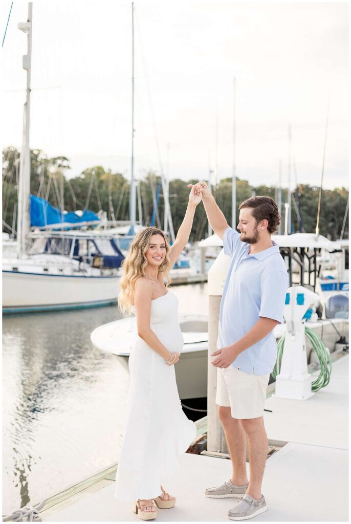 Couple stands on the dock of a marina dancing as the husband, who wears a blue polo and white shorts, looks at his wife who is wearing a white, strapless dress and twirls under his arm as she looks at the camera of Savannah maternity photographer, Wisp + Willow Photography Co.