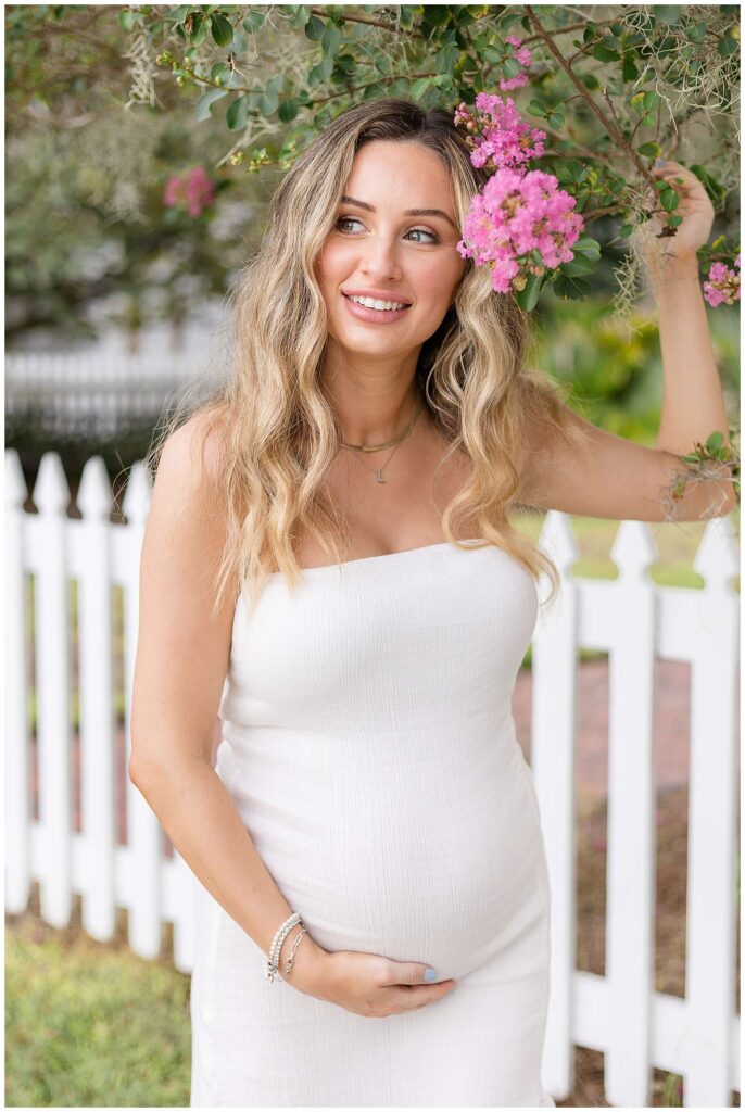 Pregnant mom-to-be stands in front of a white picket fence wearing a strapless, tight, white dress and holds the branch of a crape myrtle and smiles off into the distance during her maternity session.