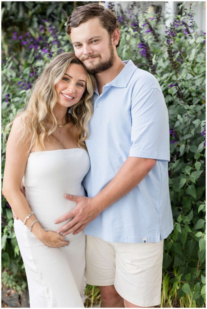 Expecting couple stand in front of a butterfly bush as they look at the camera of photographer, Wisp + Willow Photography Co.  and smile during their Savannah maternity session.