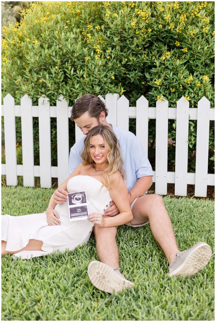 Savannah maternity photographer, Wisp + Willow Photography Co., capture a moment of a husband sitting in the grass looking down at his wife's belly who is leaning back in his lap, holding their ultrasound and smiling at the camera.