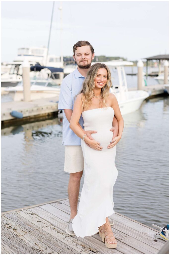 Pregnant wife, who is wearing a strapless, white, tight dress, stands on a marina dock with her husband standing behind her holding her belly as they both look at the camera and smile.