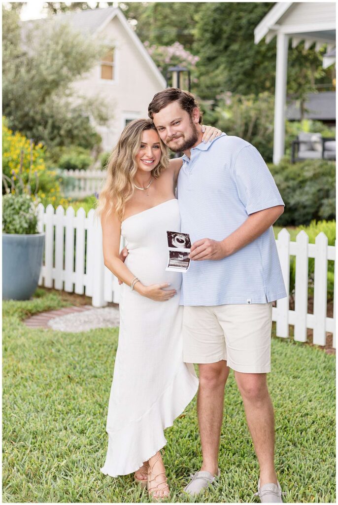 Expecting parents stand together in front of a white picket fence on green, lush grass coming cheek to cheek while holding their ultrasound picture of their baby during their Savannah maternity session.