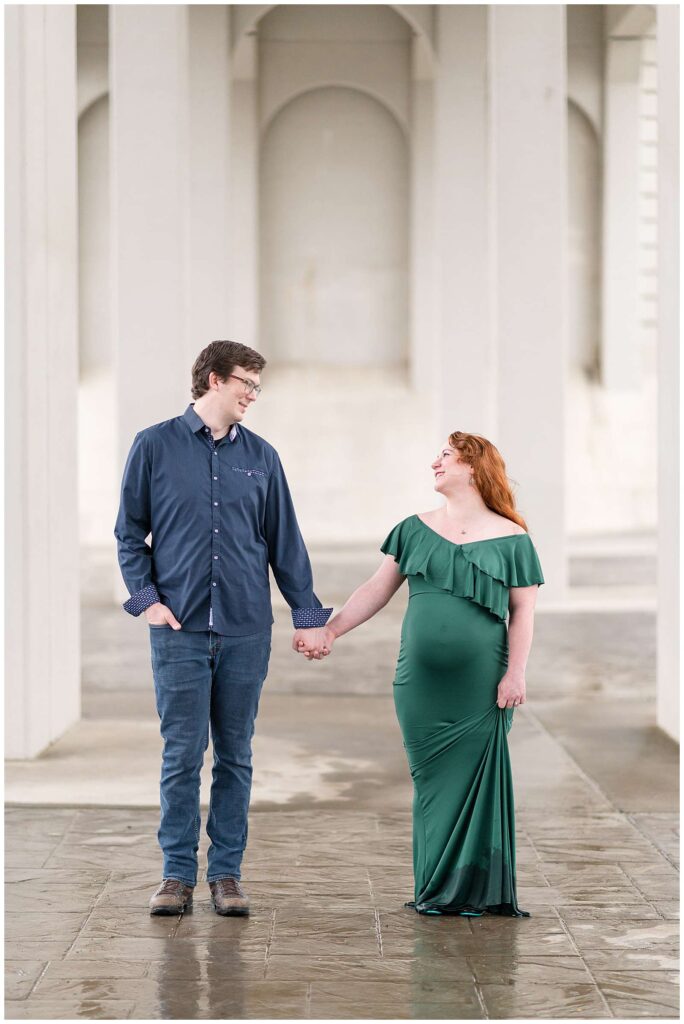 Franklin maternity photographer, Wisp + Willow Photography Co., take a picture of a couple standing slightly apart with their bodies facing the camera and they hold hands and look at each other smiling.  Pregnant mom-to- wears an emerald green, fitted dress with ruffles at the top and her husband wears a navy, button down shirt with jeans.