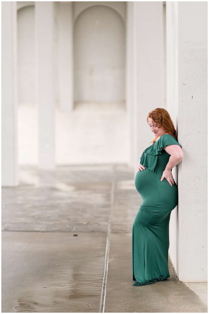 Tennesse maternity photographer, Wisp + Willow Photography Co., take a picture of a pregnant momma standing up against a square column underneath a bridge.  She wears a fitted, green, long dress with ruffles at the top and she looks down to her baby belly in wonder waiting the arrival of her baby.