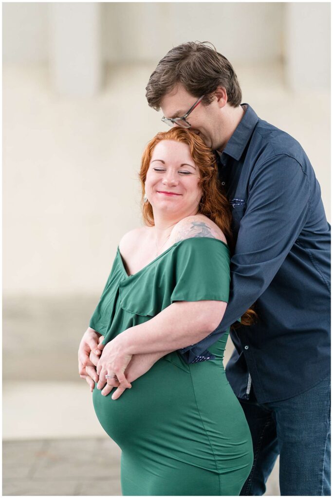 Tennessee maternity photographer captures sweet moment of husband bear hugging his wife from behind putting his hands on her belly as she wraps her hands on top of his and smiles while closing her eyes.