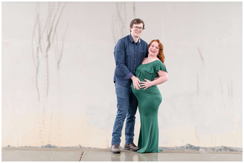 Franklin, TN maternity photographer, Wisp + Willow Photography photograph a couple in front of a neutral wall standing under a bridge as the smile at the camera.  The husband, who is wearing a navy, button down shirt and jeans puts one hand on his wife's belly as she stands belly to belly with him wearing a green, fitted, long dress and has red hair.