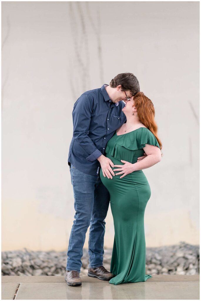 Franklin, TN maternity photographer takes a picture of expecting couple as they stand belly to belly looking at each other and coming in close nose to nose.  He wears a navy button down shirt and blue jeans and she wears a green, fitted, long dress and has bright, red hair.