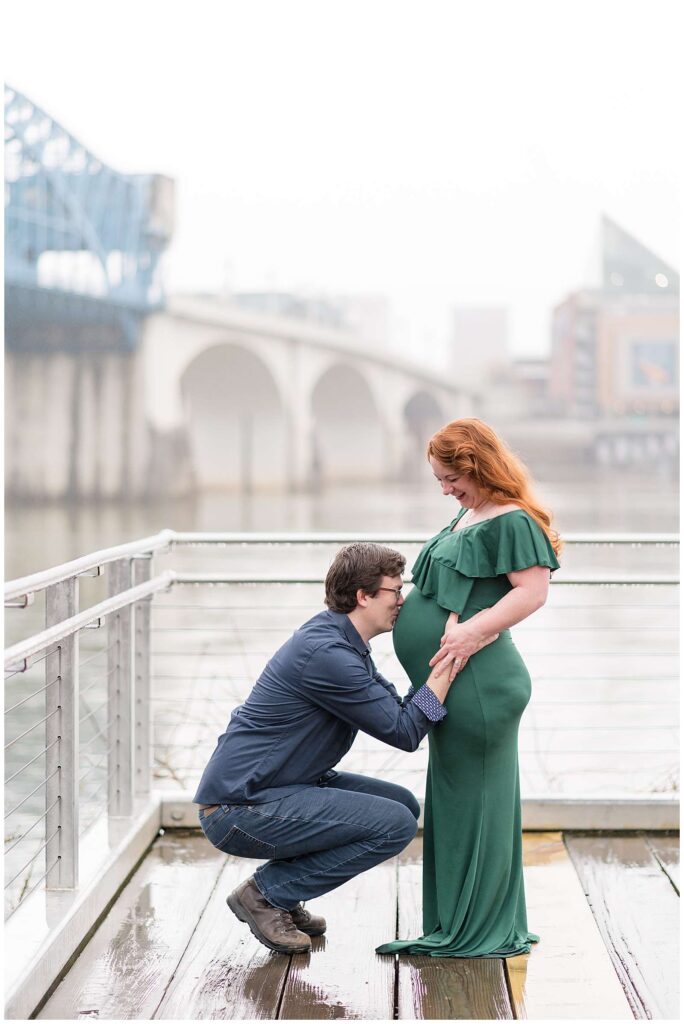 Wisp + Willow Photography Co. have a maternity photography session at Coolidge Park in Franklin, TN in the rain.  They stand on a walkway over the water with a bridge in the back as the husband squats down to kiss his wife's belly with their expecting child inside.  Preganant momma wears a green, ruffled, dress that goes down to the ground and is fitted over the belly.