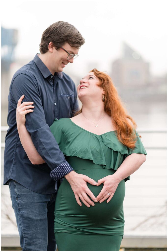 Maternity photographer, Wisp + Willow Photography Co., take a picture of an expecting couple standing in the drizzle looking up at each other.  The both have a hand on the baby belly as they make a heart together with their hands.
