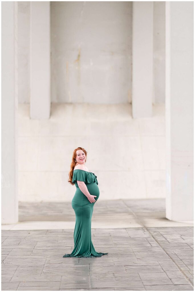 Chattanooga maternity photographer, Wisp + Willow Photography Co., take a picture of mom-to-be who has bright, red hair and wears an off the shoulder green dress with ruffles at the top and fitted down to the ground.  She stands with her body facing the right as she looks over and smiles at the camera.