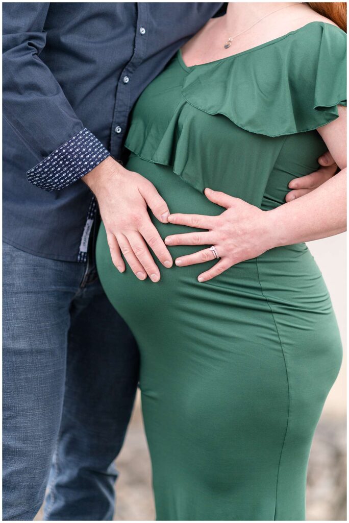 Close up image of expecting couple shows both of their hands on the pregnant belly.  Mom-to-be wears a green ruffled, fitted dress and her husband wears a navy button down shirt and dark, blue jeans.