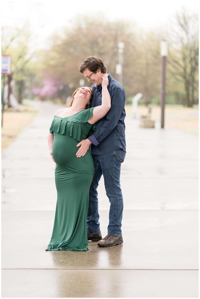 Wisp + Willow Photography Co, Chattanooga maternity photographer, captures couple standing on a sidewalk in Collidge Park in TN.  The mom-to-be wears a dark green, long, fitted dress as she leans back towards her husband holding his neck as he looks down and they laugh together.
