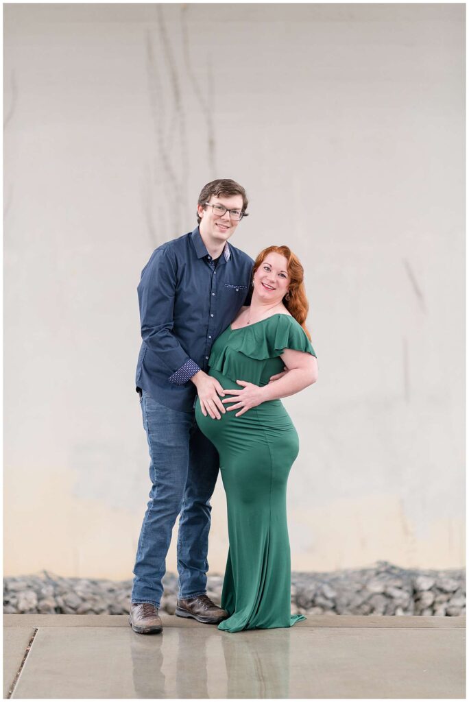Chattanooga maternity photographer has couple under bridge during a rainy session.  Mom to be, wears a long, fitted green dress with a ruffled top and her husband wears a navy, button down shirt and jeans holding his wife's belly as they both look at the camera.